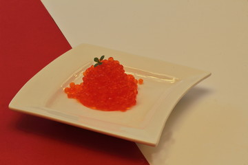 Red and white. Red salted salmon roe caviar with lettuce cress on white porcelain plate of unusual shape on contrasting white and red background. Minimalism style.