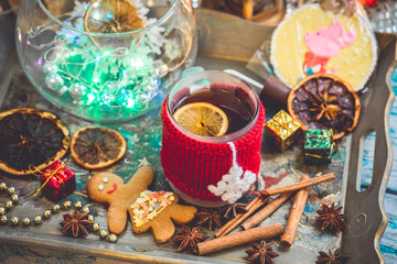 New Year's still life with mug of tea with knitted cup cover, cookies, chocolate, dried fruit and seasonings with lights and bokeh. Photo toned and with vignette