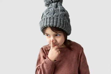 Closeup horizontal portrait of cute little girl in the winter warm gray hat, wearing sweater touching her nose, posing on a white studio background.