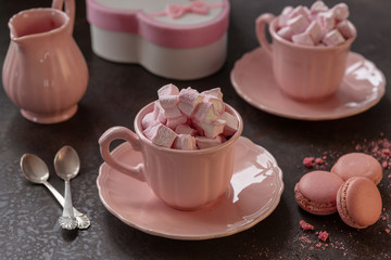 Two pink cups with pink heart-shaped marshmallows, a gift in a pink box and pink macarons. Romantic breakfast for two. Close-up, selective focus.