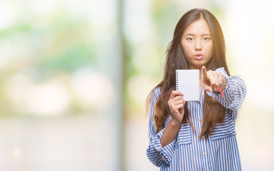 Young asian woman holding notebook over isolated background pointing with finger to the camera and to you, hand sign, positive and confident gesture from the front