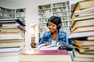 Pretty young african woman sitting and listening to music while she is studying her books in modern university library. Education, studying concept