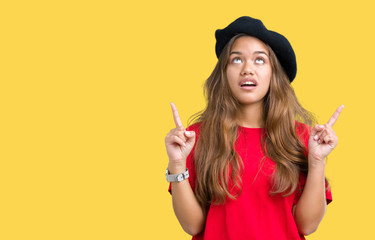 Young beautiful brunette fashion woman wearing red t-shirt and black beret over isolated background amazed and surprised looking up and pointing with fingers and raised arms.