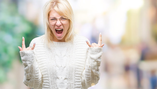 Young beautiful blonde woman wearing winter sweater and glasses over isolated background shouting with crazy expression doing rock symbol with hands up. Music star. Heavy concept.