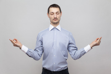 Portrait of calm handsome bristle businessman in classic light blue shirt standing in yoga pose with closed eyes and meditating. indoor studio shot, isolated on grey background copyspace.