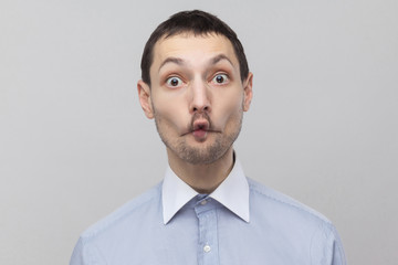 Portrait of funny handsome bristle businessman in classic blue shirt standing looking at camera with surprised face, big eyes and fish lips . indoor studio shot, isolated on grey background copyspace.