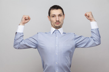 I am strong. Portrait of serious proud handsome bristle businessman in classic blue shirt standing and looking at camera with raised arms. indoor studio shot, isolated on grey background copyspace.