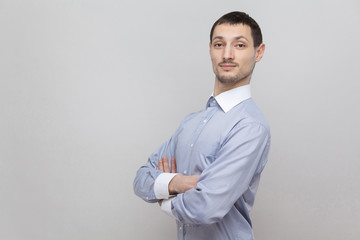Portrait of handsome bristle businessman in classic light blue shirt standing, crossed hands and looking at camera with confident face. indoor studio shot, isolated on grey background copyspace.