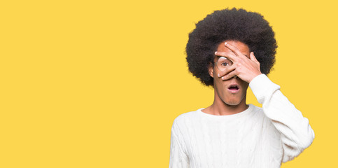 Young african american man with afro hair wearing glasses peeking in shock covering face and eyes with hand, looking through fingers with embarrassed expression.