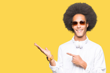 Young african american man with afro hair wearing sunglasses and headphones amazed and smiling to the camera while presenting with hand and pointing with finger.