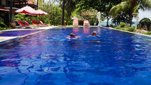 man and woman swims in swimmingpool in luxury hotel. couple leisure in the pool luxury hotel with swimming pool, sun beds, palm trees by sea. tropical resort with pool. Travel concept.