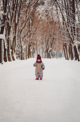 Portrait of a cute baby dressed in a gray jacket and a red hat that walks through the snow covered snow park. She smiles one in the photo during the snowfall