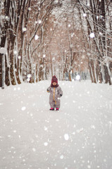 Portrait of a cute baby dressed in a gray jacket and a red hat that walks through the snow covered snow park. She smiles one in the photo during the snowfall