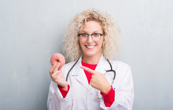 Young blonde dietist doctor woman over grunge grey wall holding donut very happy pointing with hand and finger