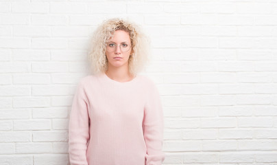 Young blonde woman with curly hair over white brick wall with serious expression on face. Simple and natural looking at the camera.