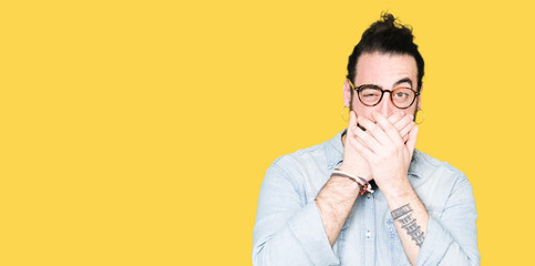 Young hipster man with long hair and beard wearing glasses shocked covering mouth with hands for mistake. Secret concept.