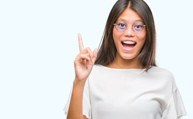 Young asian woman wearing glasses over isolated background pointing finger up with successful idea. Exited and happy. Number one.