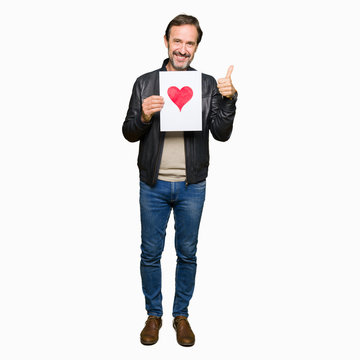 Handsome middle age man holding paper with red heart happy with big smile doing ok sign, thumb up with fingers, excellent sign
