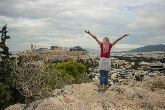 travel girl back to camera in freedom pose with hand up on top of rock with Acropolis world heritage touristic site in Athens city - capital of Greece