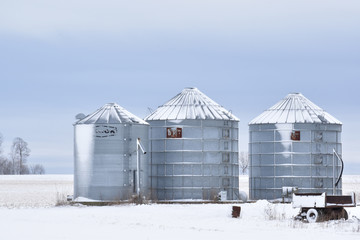 Three silver metal grain feed storage silos covered in snow on a farm in winter in the country © Jennifer