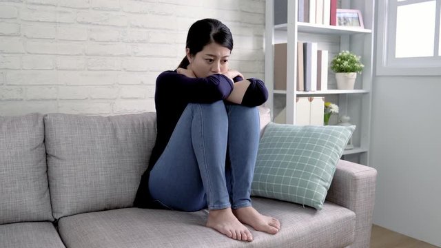 Depressed young asian woman sitting on couch at home in bright living room. Unhappy lonely chinese lady in sofa hiding her face in her cross arms and legs. housewife feel unsatisfied about life