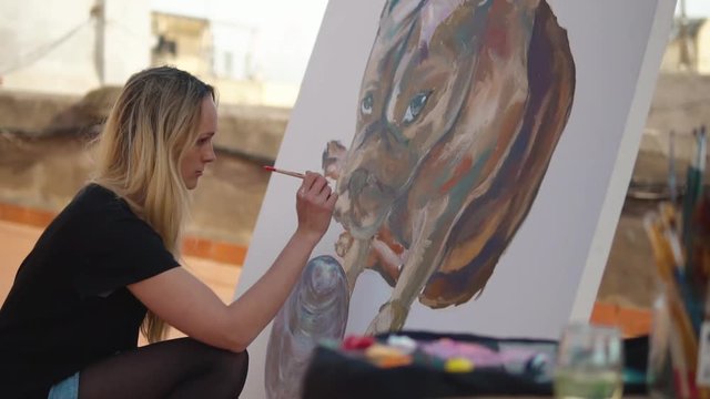 Young woman paint artist drawing at home roof. Picture of bulldog on big canvas. Outdoors slowmotion art