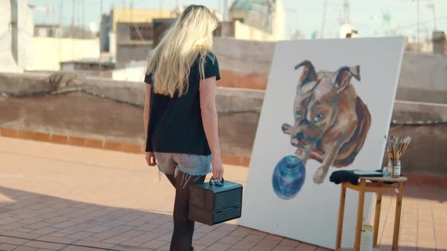 Young woman paint artist coming for drawing at home roof. Picture of bulldog on big canvas. Outdoors slowmotion art