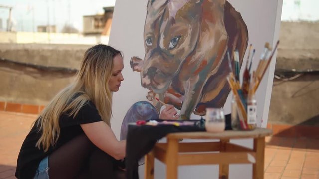 Young woman paint artist drawing at home roof. Picture of bulldog on big canvas. Outdoors slowmotion art