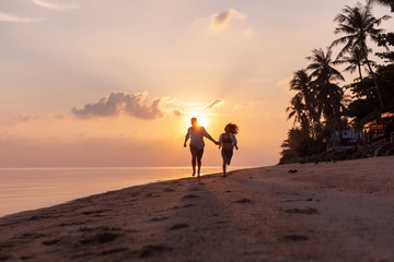 Beautiful happy young couple in love runs along the beach along the sand by the sea at sunset during a honeymoon holiday vacation