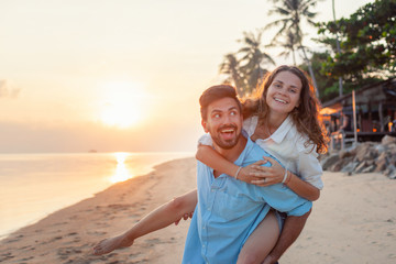Beautiful young happy couple in love having fun on the beach at sunset during the honeymoon vacation travel, the guy carries the girl on yourself