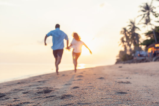 Young couple running along the sandy seashore in the rays of sunset, blurred image perfect background for travel agencies, honeymoon vacation travel in the tropics