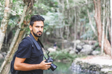 Handsome young stylish male photographer in black t-shirt and sunglasses is engaged in trekking in the green jungle.