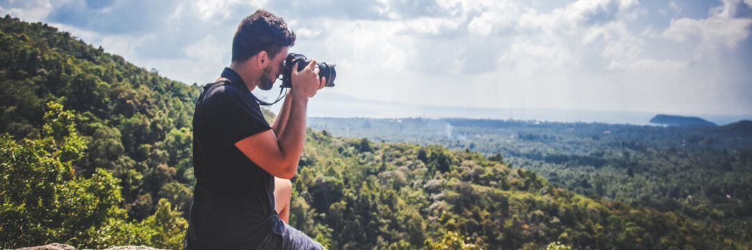 Handsome young stylish male photographer of mixed race in a black T-shirt with a camera in hand on top of a mountain with a view