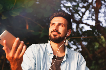Young attractive man of mixed race guy Latino with a beard listening to music on headphones, outdoors