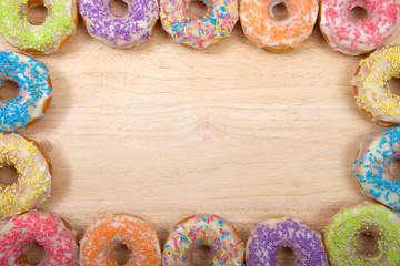 Flat lay view of many Frosted donuts with candy sprinkles arranged in a border frame on a light wood table with copy space.