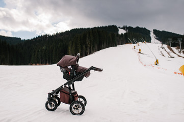 Baby stroller is on snow in the Carpathian mountains, outdoors. On background of the forest and ski slopes with lifts. Close up. Winter nature. Hoist in the mountains. Cannons that make snow.