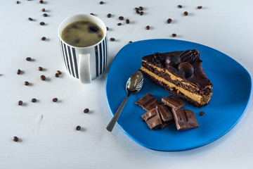 A piece of chocolate cake, and pieces of chocolate on a blue plate, next to a cup of coffee, on a light wooden background.