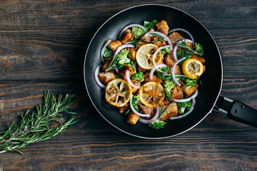 Fried pork with broccoli, lemon and rosemary in a pan on a dark wooden table top view