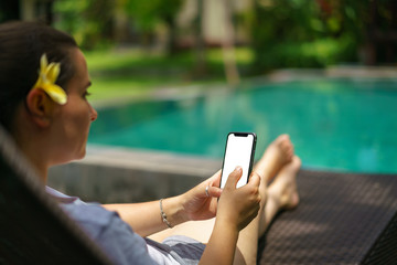 Woman sitting in deck chair tropical pool holding the black smart phone with blank screen and...