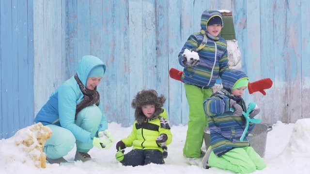 Three happy children in winter warm clothes make snowmen and play with them. White Snowman dressed in scarf, tin bucket with blue eyes and carrot nose against background of wooden fence. Christmas Eve