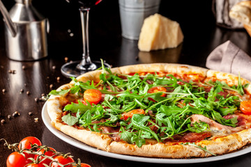 Obraz na płótnie Canvas The concept of Italian cuisine. Pizza with sausage salami, bacon, cherry tomatoes and arugula. Next to the table is a glass of red wine. Beautiful serving dishes in the restaurant.