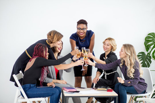 Positive, stylish, attractive, cheerful, business ladies in casual outfits celebrating event clinking glasses with champagne looking at birthday girl, sitting in office lounge zone