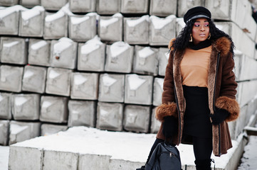 African american woman in sheepskin coat and cap posed at winter day against snowy stone background.