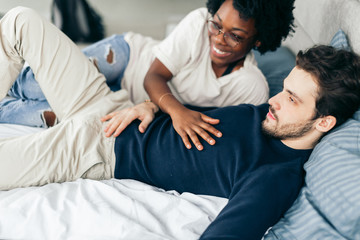 Obraz premium Passionate young african woman seducing her boyfriend in bed while caucasian handsome man watching TV