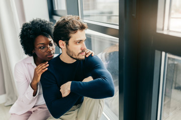 Loving thoughtful interracial couple sitting on the sill and looking at the large window in their hired flat. Caucasian guy wears a dark poolover, african girl wears pink shirt. - Powered by Adobe