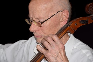 Alter, weißer Mann mit weißem Oberhemd spielt konzetriert Cello vor dunklem Hintergrund