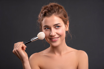 Obraz premium Beautiful brunette caucasian young woman prepare herself, applying powder on her cheeks with a brush. Clean, fresh, natural, flawless skin. Soft smile on her face. Close up on a neutral background