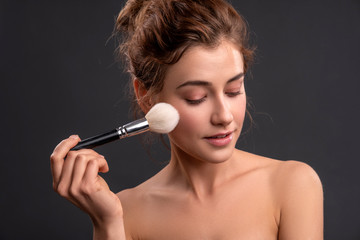 Obraz premium Beautiful brunette caucasian young woman prepare herself, applying powder on her cheeks with a brush. Clean, fresh, natural, flawless skin. Soft smile on her face. Close up on a neutral background