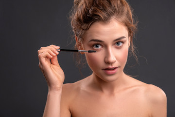 Obraz premium Beautiful brunette caucasian young woman prepare herself, applying mascara on her eyes with a brush. Clean, fresh, natural, flawless skin. Soft smile on her face. Close up on a neutral background