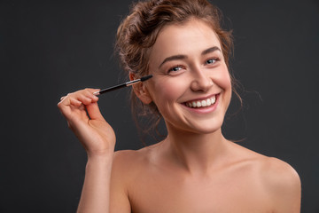 Fototapeta premium Beautiful brunette caucasian young woman prepare herself, applying mascara on her eyes with a brush. Clean, fresh, natural, flawless skin. Soft smile on her face. Close up on a neutral background